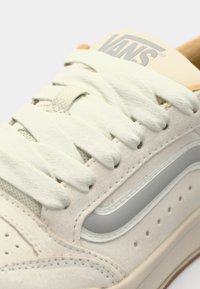 Vans-sneakers med lys beige semsket skinn, tonale snørebånd, polstret tunge med logo, og en grå side stripe med perforeringer langs tåen.