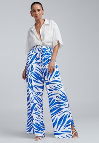 Pantalons à jambe large dans un motif abstrait bleu et blanc, avec une taille élastique et des fentes sur les côtés. Portés avec une chemise blanche à col.