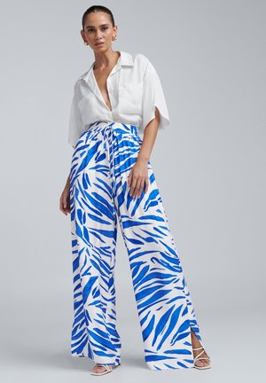 Pantalons à jambe large dans un motif abstrait bleu et blanc, avec une taille élastique et des fentes sur les côtés. Portés avec une chemise blanche à col.