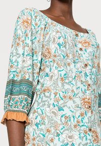 Ljusblå blommig klänning med orange och vita blommor, trekvartslånga ärmar med volangmuddar och träknappar framtill.