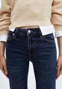 Jean en denim bleu foncé taille haute, avec des surpiqûres orange contrastantes et une fermeture à bouton argenté. Associé à un pull beige.