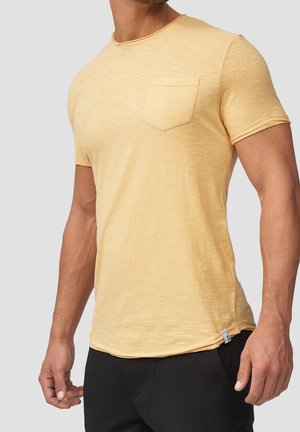 Maglietta gialla a maniche corte con scollo rotondo, dotata di taschino sul petto e tessuto morbido e strutturato. È abbinata a pantaloni neri.