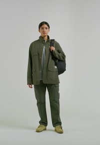 Olivgrön, lätt jacka med två bröstfickor, matchad med cargo-byxor. Grå t-shirt under och svart ryggsäck. Beige sneakers.