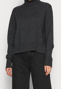 Un pull col roulé gris foncé en tricot texturé, avec un ourlet court et des poignets côtelés, associé à un pantalon noir à jambes larges.