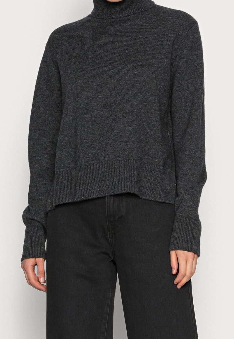 Un pull col roulé gris foncé en tricot texturé, avec un ourlet court et des poignets côtelés, associé à un pantalon noir à jambes larges.