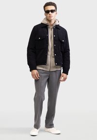 Black corduroy jacket with front pockets, beige hoodie underneath, grey tailored pants, and white sneakers. Model wearing sunglasses.