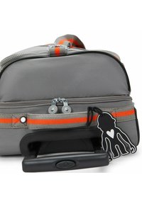 Bolsa de viaje de tejido suave en color gris con una franja de acento naranja, doble cremallera, mango telescópico y un llavero de mono negro adjunto.