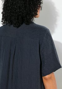 Tmavě modrý tkaný top s krátkými rukávy, s texturovaným povrchem a záhybem na zadní straně pro zvýraznění detailu.