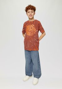 Jongen in een rood-oranje tie-dye t-shirt met vetgele tekst, gecombineerd met losse blauwe jeans en witte sneakers. Neutrale achtergrond.