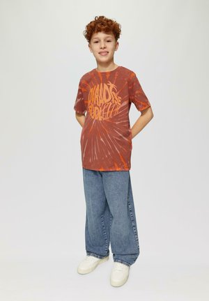 Junge trägt ein rot-orangefarbenes Tie-Dye-T-Shirt mit fettem gelben Text, kombiniert mit lockeren blauen Jeans und weißen Turnschuhen. Neutraler Hintergrund.