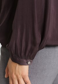 Langærmet bluse i dyb plommefarve med silkeagtig tekstur, sammenfoldede manchetter og sølvknapdetalje, parret med lys grå stof.