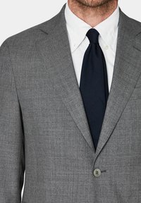 Giacca da abito grigia in tessuto con chiusura a un bottone e reverse a dente di cane, abbinata a una camicia bianca e una cravatta blu navy.