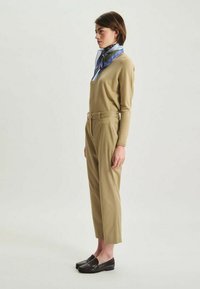 Pull léger marron, pantalon beige taille haute avec ceinture, mocassins noirs, et une écharpe bleue à motifs. Textures lisses et design ajusté.