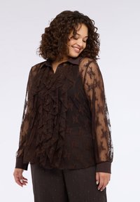 Blusa trasparente marrone scuro con volant sovrapposti, motivo floreale e maniche lunghe. Presenta un colletto appuntito e dettagli con bottoni sui polsini.