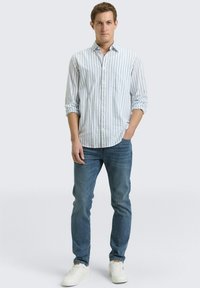 Man staat met één hand in zijn zak, draagt een wit gestreept shirt met lange mouwen, blauwe spijkerbroek en witte sneakers tegen een effen achtergrond.