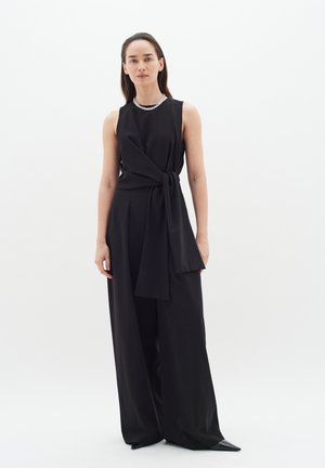 Un salopette nera senza maniche con design avvolgente e dettaglio da annodare sul davanti. Il tessuto liscio, le gambe ampie e le scarpe a punta accentuano il look.