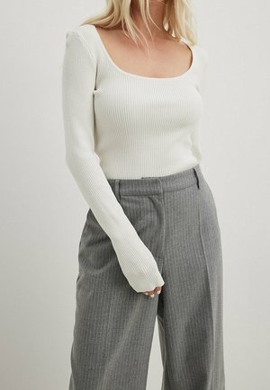 Frau trägt ein weißes, geripptes Langarmshirt mit quadratischem Ausschnitt und hoch taillierte graue Nadelstreifenhose.