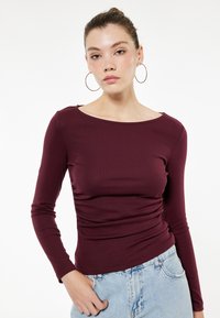 Burgundin pitkähihaisen pelti, jossa on ribbed-rakenne, pyöreä kaula-aukko ja keräsomistus yhdellä puolella, yhdistettynä vaaleisiin farkkuihin.