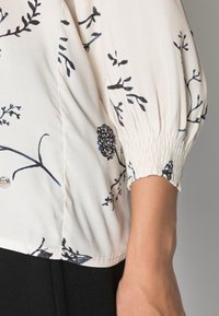 Crème blouse met marineblauwe bloemenprint, geraffineerde mouwen en smockmanchetten. Heeft een ronde halslijn en een subtiele textuurstof.