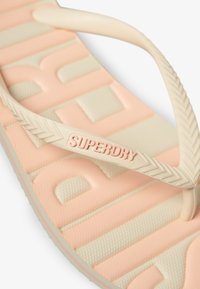 Tongs avec une semelle en caoutchouc rose doux, une sangle beige arborant le logo "SUPERDRY", surface texturée avec des lettres en relief et des motifs géométriques.