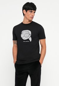 Čierne bavlnené tričko s grafickým vzorom stylizovanej tváre v slnečných okuliaroch pokryté strieborným trblietaním, pričom pod ním je vytlačené "KARL LAGERFELD".