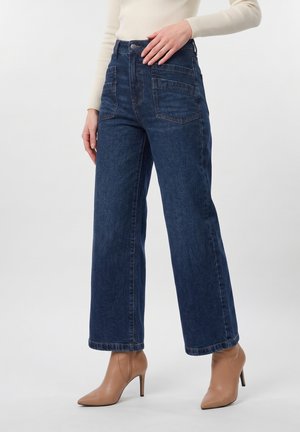Kvinde iført mørkeblå, bredbenede jeans med høj talje, store forlommer og beige ankelstøvler med høj hæl og spids tå.