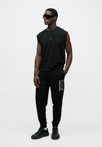 Črna brezrokavnik s logotipom, kombinirana s črnimi trenirkami z logotipom ob strani in črnimi supergami; gladek material z udobnim krojem.