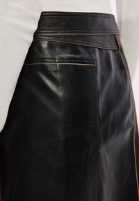 Μαύρη δερμάτινη φούστα με λεία υφή, αντίθεση ραφών κατά μήκος των ραφών και εφαρμοστή ζώνη. Ορατή λεπτομέρεια με πλαϊνές τσέπες.