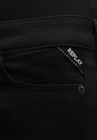 Κοντινό πλάνο στην τσέπη μαύρου τζιν με μαύρη και άσπρη ετικέτα "Replay" ραμμένη στη γωνία πάνω από έναν πριτσίνι.