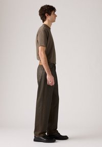 Mlad moški stoji bočno, oblečen v rjav majico, temno rjave hlače, črne čevlje in prstan na desni roki.