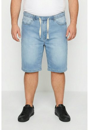 Uomo che indossa pantaloncini di jeans azzurri chiari con coulisse in vita, sneakers nere e una maglietta bianca tagliata in vita, in piedi su sfondo bianco.