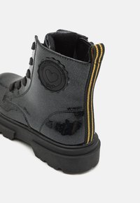 Bottine noire en matériau brillant avec une finition texturée, mettant en avant un accent à rayures jaunes et un patch logo rond sur le côté.