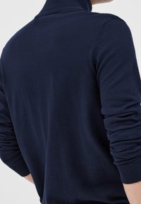 Man som bär en långärmad marinblå tröja med ribbade muddar och en något upphöjd krage, sedd från ryggen.