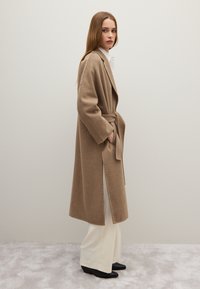 Cappotto lungo beige in lana con cintura, maniche aderenti, spacchi laterali e tasche, indossato su una camicia bianca e pantaloni a gamba larga color crema. Scarpe nere.