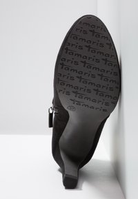 Bottine noire à talon, inclinée pour montrer la semelle avec le motif de texte "tamaris" et la taille 37 indiquée.