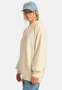 Beige oversized hoodie met een voorzak en een trekkoord, voorzien van geborduurde tekst "PLAYING"; gecombineerd met een lichtblauwe denim pet en jeans.