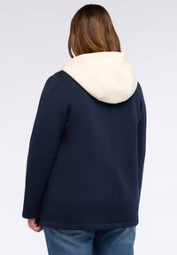 Giacca a cappuccio blu navy con interno crema, realizzata in tessuto morbido. Presenta una vestibilità comoda e un design semplice, con una cucitura pulita lungo la schiena.