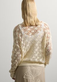 Mujer con cabello rubio hasta los hombros que lleva un suéter de punto abierto crema y una falda beige con un gran diseño de hoja bordado, mostrada desde la espalda.