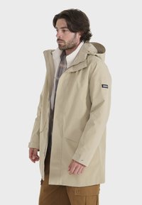 Beige vandtæt jakke med hætte, frontlynlås og knaplukninger. Har et blødt indre for og et logo-patch på venstre ærme.