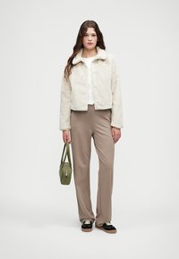 Krem kaput od lažnog krzna s ovratnikom, nosi se preko bijele košulje. Bež krojene hlače, zeleni ručni torbica i crne cipele s naglascima upotpunjuju odjevnu kombinaciju.