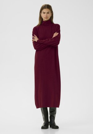 Rochie burgundy cu guler înalt, realizată dintr-un material tricotat moale, având mâneci lungi și o siluetă dreaptă, asortată cu cizme negre până la genunchi.