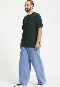 Ciemnozielony oversize'owy t-shirt z krótkimi rękawami, zestawiony z jasnoniebieskimi jeansami o szerokich nogawkach. Dopełniony czarnymi niskimi trampkami.