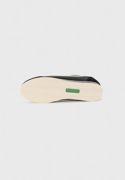 Black sneaker with a textured upper, cream rubber sole, and green brand label. Features a herringbone tread pattern for grip.