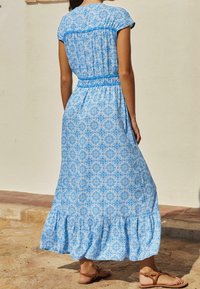 Maxi sukienka z jasnoniebieskiego materiału, zdobiona kwiatowym wzorem, z krótkimi rękawami, gumką w talii i falbanistym dołem. Całość dopełniają płaskie sandały.