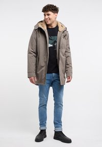 Brun, texturerad parka med en huva foder av syntetpäls, med dragkedja och knappstängning, bärs över en svart t-shirt med grafik och blå jeans.