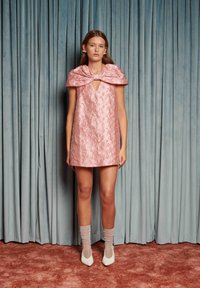 Kvinne står foran blå gardiner iført en rosa teksturert minikjole med en stor sløyfe krage, gjennomsiktige glitrende sokker og hvite hæler.