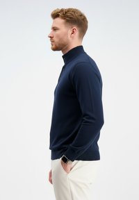 Vyras, dėvintis tamsiai mėlyną megztinį su sagomis ir kreminiais kelnėmis, stovintis profiliu su viena ranka kišenėje priešais vienspalvį foną.