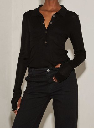 Person wearing black long-sleeve henley shirt with button placket and black high-waisted pants, one hand in pocket, against plain background.