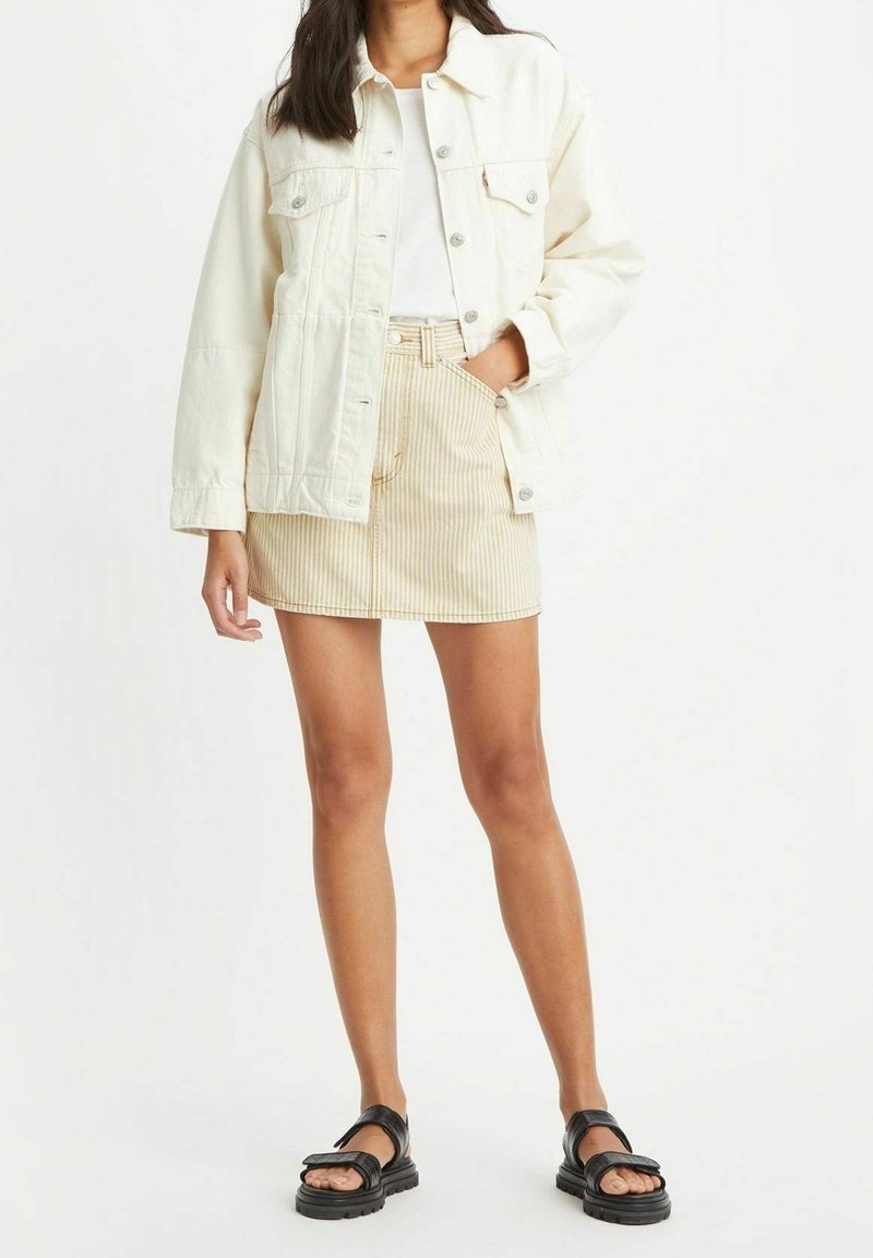 Femme portant une veste en jean blanche, un haut blanc, une mini-jupe beige rayée et des sandales noires, debout avec une main dans la poche de la jupe.