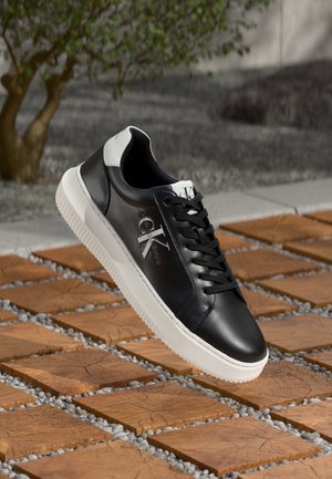 Schwarze Leder-Sneaker mit weißer Gummisohle, featuring Schnürdesign und Logodetail an der Seite. Der Hintergrund umfasst Stein- und Holztexturen.